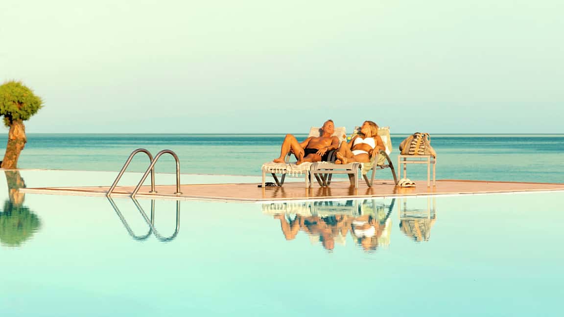 Kaksi henkilöä makaa aurinkotuoleilla uima-altaan äärellä, näkymä merelle. (Tekoälyn luoma teksti)