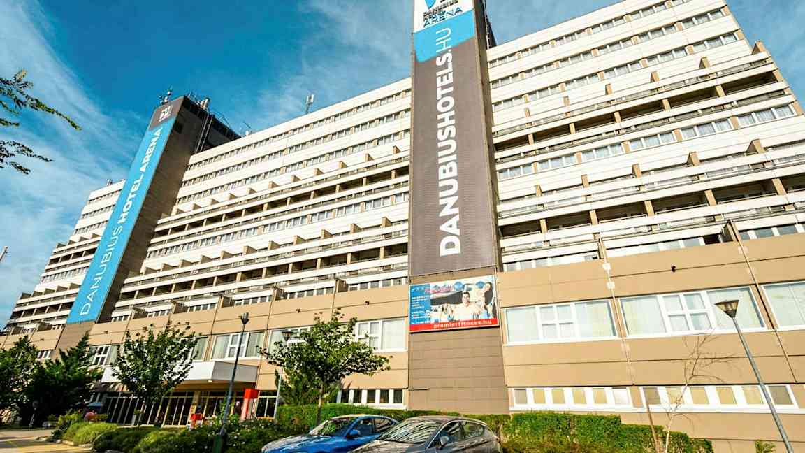 Suuri hotellirakennus, jossa on useita kerroksia ja näkyvässä kyltissä lukee ”Danubius Hotel Arena”. Rakennuksen edessä on pysäköityjä autoja ja joitakin pieniä puita. (Tekoälyn luoma teksti)