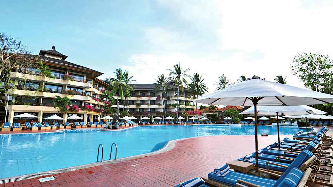 Kuva ulkouima-altaasta, jota ympäröivät aurinkotuolit ja aurinkovarjot, taustalla monikerroksinen hotelli ja palmuja. (Tekoälyn luoma teksti)