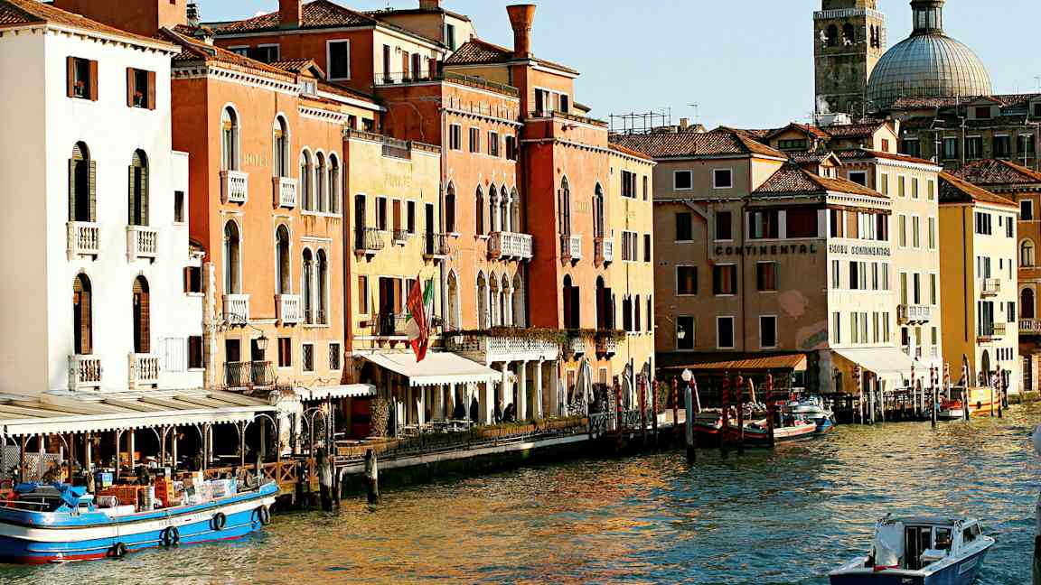 Kuva Venetsian kanavasta, jossa on värikkäitä rakennuksia, veneitä ja kupolimainen kirkko taustalla. (Tekoälyn luoma teksti)