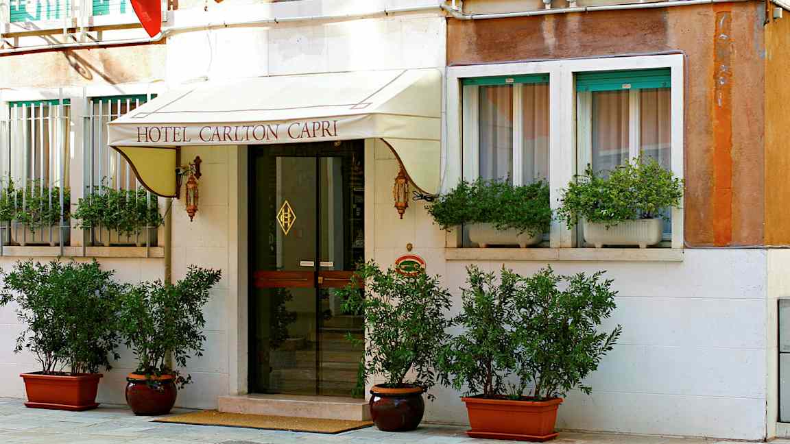 Hotel Carlton Caprin sisäänkäynti kyltti, lasiovi ja kasveja. (Tekoälyn luoma teksti)