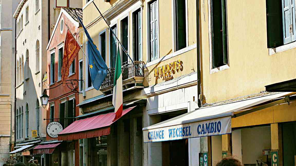 Rakennusten julkisivu, italialainen lippu, eurooppalainen lippu ja kyltit, jotka osoittavat hotellin ja vaihdon. (Tekoälyn luoma teksti)