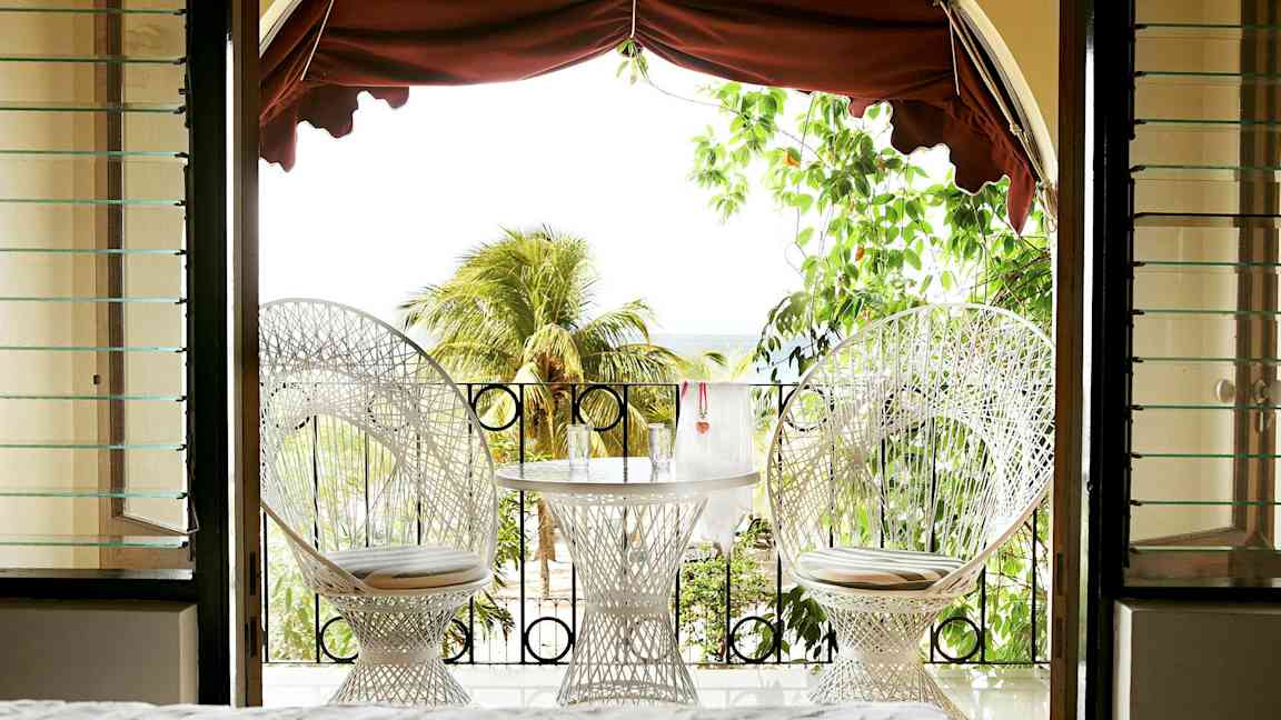 Näkymä huoneesta terassille, jossa on kaksi valkoista tuolia ja pöytä laseineen, ympäröitynä vehreydellä ja palmupuilla. (Tekoälyn luoma teksti)
