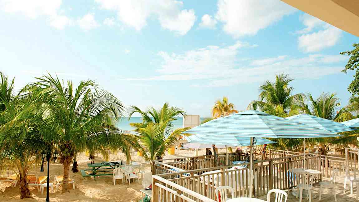 Kuva rannasta, jossa on palmuja, pöytiä ja tuoleja sekä aurinkovarjoja. Taivas on selkeä ja siellä on muutamia pilviä. (Tekoälyn luoma teksti)