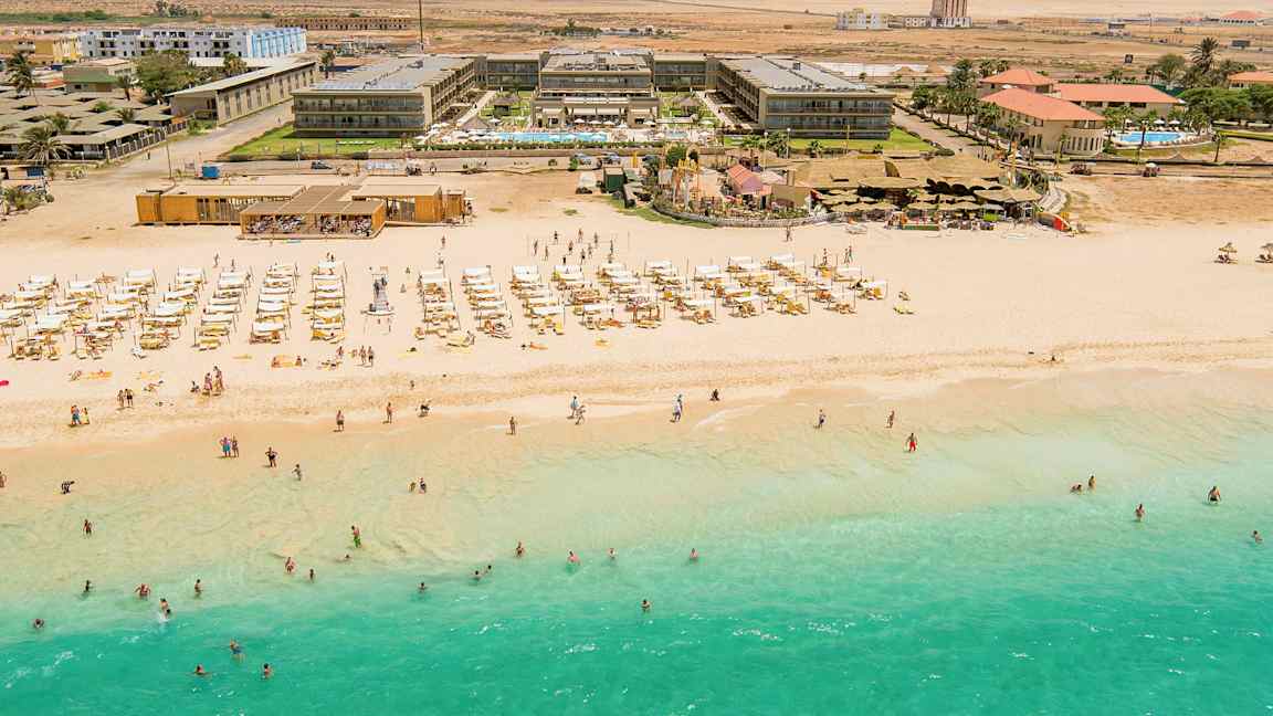 Ilmakuva rannasta, jossa on turkoosia vettä, aurinkotuoleja ja aurinkovarjoja, sekä hotellirakennus taustalla. (Tekoälyn luoma teksti)