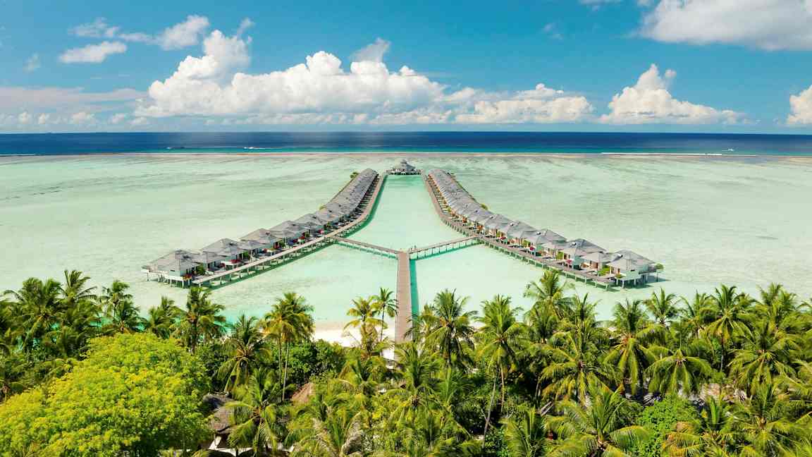 Kuvassa veden päällä olevia bungalow-taloja yhdessä kylämuodossa, kujan ympäröimänä palmujen sekä sinisen meren ja taivaan reunamilla. (Tekoälyn luoma teksti)