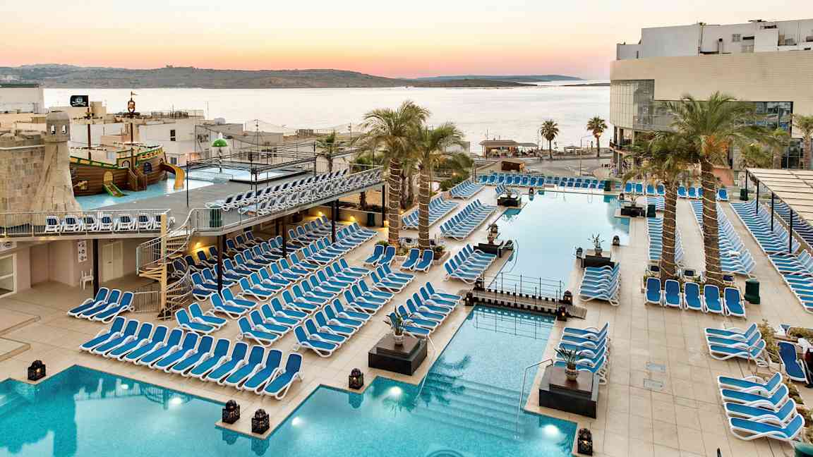 Suuri allasalue aurinkotuoleilla, palmuilla ja näköalalla merelle auringonlaskun aikaan. (Tekoälyn luoma teksti)