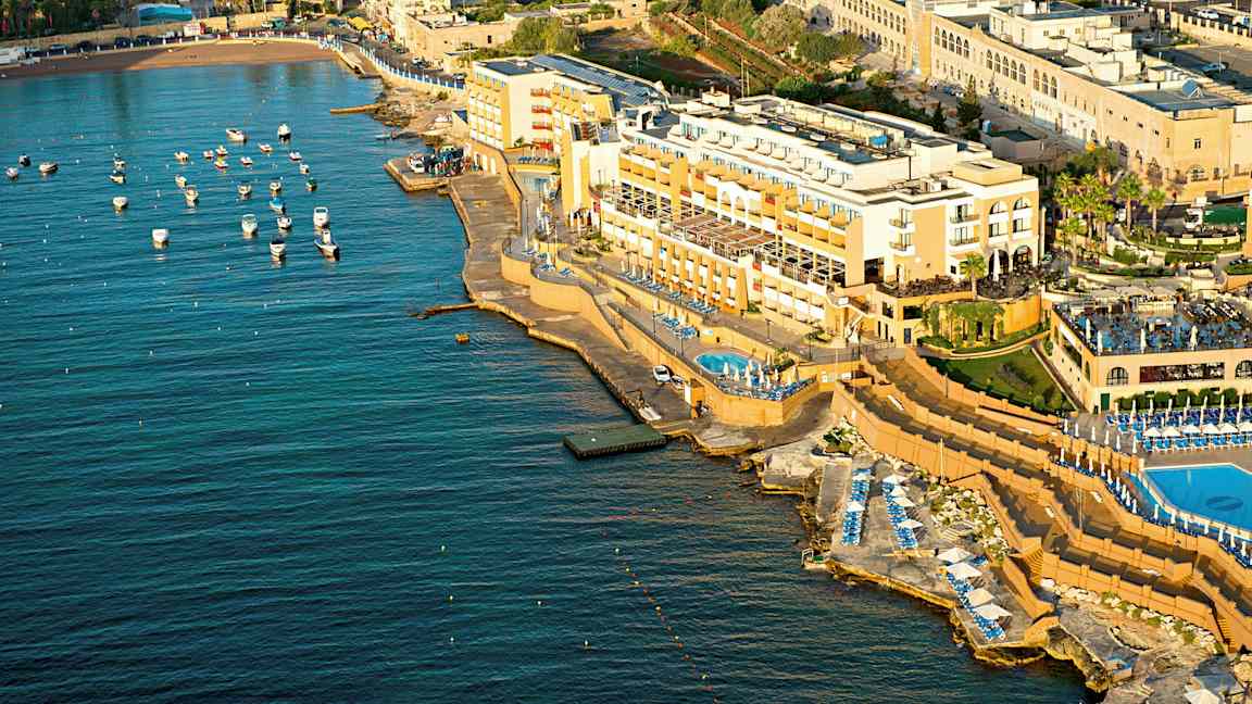 Ilmakuva rannikon hotellista, jossa on uima-altaita ja aurinkotuoleja meren äärellä. Pieniä veneitä on kiinnitetty veteen. (Tekoälyn luoma teksti)