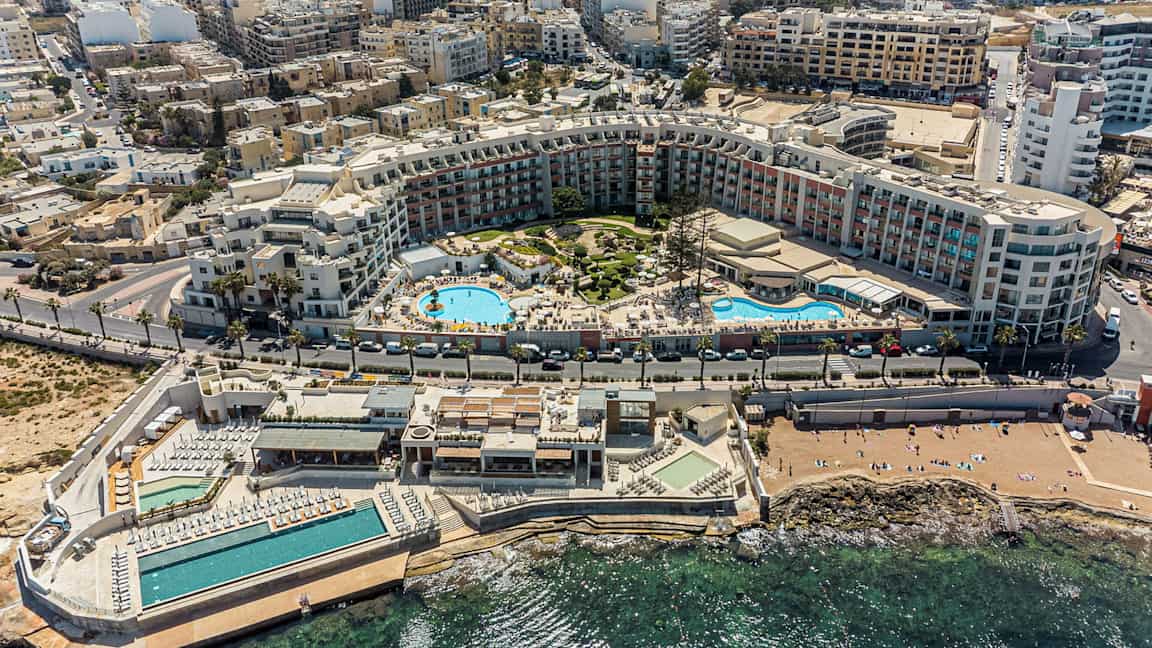 Kuvassa on laaja lomakeskus, jossa on uima-altaita, auringonottoalueita ja ympäröiviä rakennuksia rannikolla. (Tekoälyn luoma teksti)