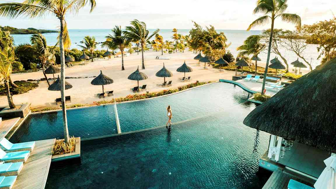 Trooppinen ranta, jolla on palmuja ja aurinkovarjoja meren äärellä, edessä infinity-allas. (Tekoälyn luoma teksti)