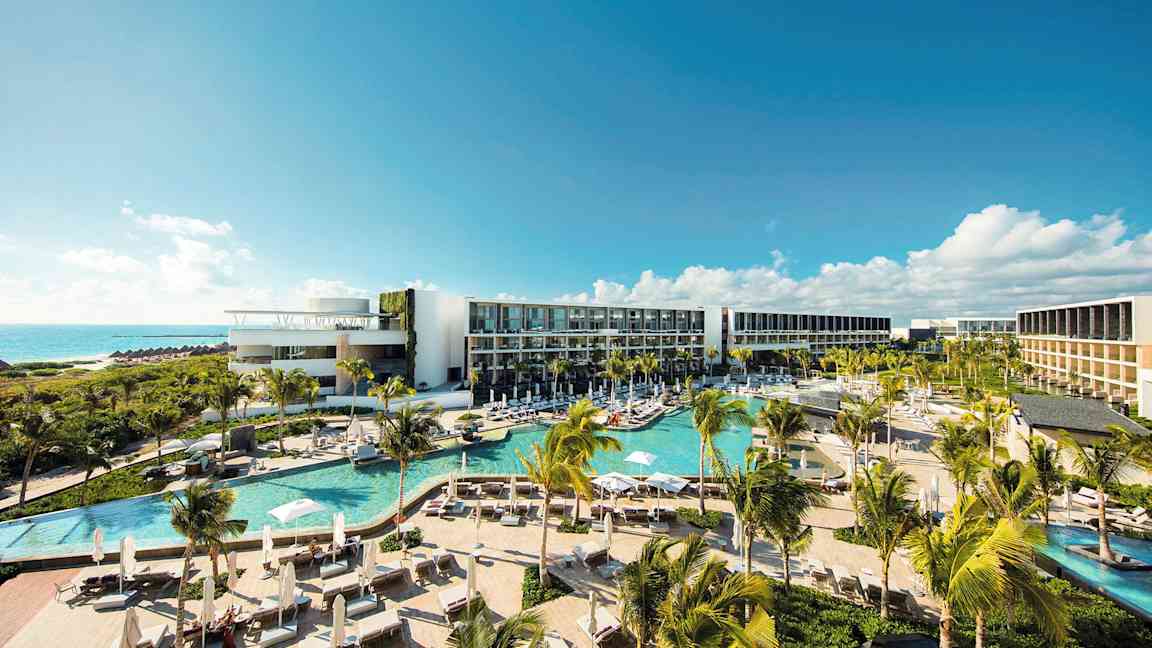 Nykyaikainen hotellialue, jossa on useita uima-altaita, vehreää kasvillisuutta ja palmuja, sijaitsee rannikolla ja tarjoaa näkymän merelle kirkkaan sinisen taivaan alla. (Tekoälyn luoma teksti)