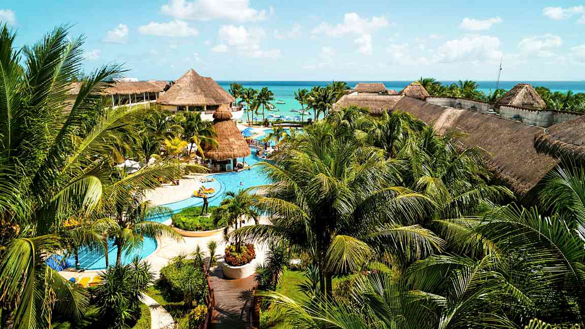 Suuri lomakeskus, jossa on uima-altaita, palmuja ja olkikattoisia rakennuksia, sijaitsee lähellä merta sinisen taivaan alla. (Tekoälyn luoma teksti)