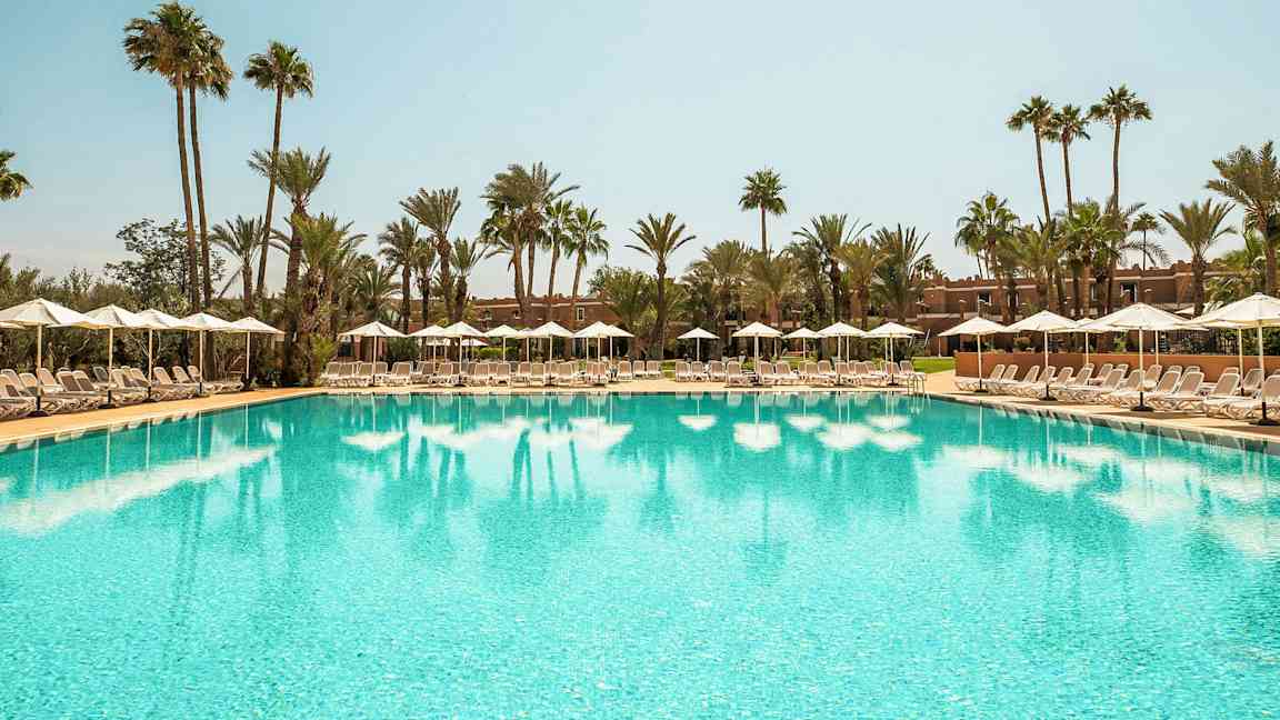 Suuri uima-allas, jonka ympärillä aurinkotuoleja ja sateenvarjoja, kirkkaan taivaan alla, taustalla palmuja. (Tekoälyn luoma teksti)