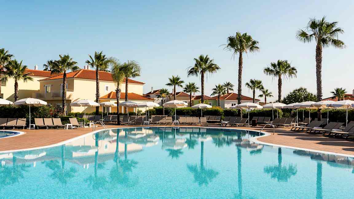 Ulkouima-allas aurinkotuoleilla ja varjoilla, ympärillä palmuja ja rakennuksia, joissa on tiilikatto. (Tekoälyn luoma teksti)