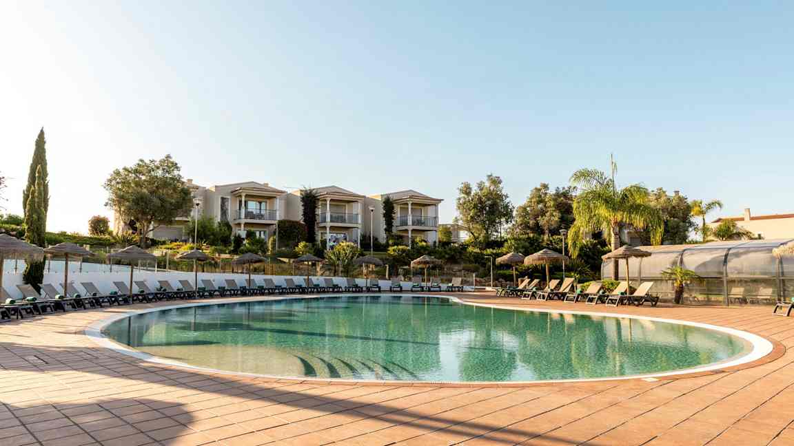 Ulkouima-allas, jossa aurinkotuoleja ja aurinkovarjoja, ympärillä vehreyttä ja rakennuksia. (Tekoälyn luoma teksti)