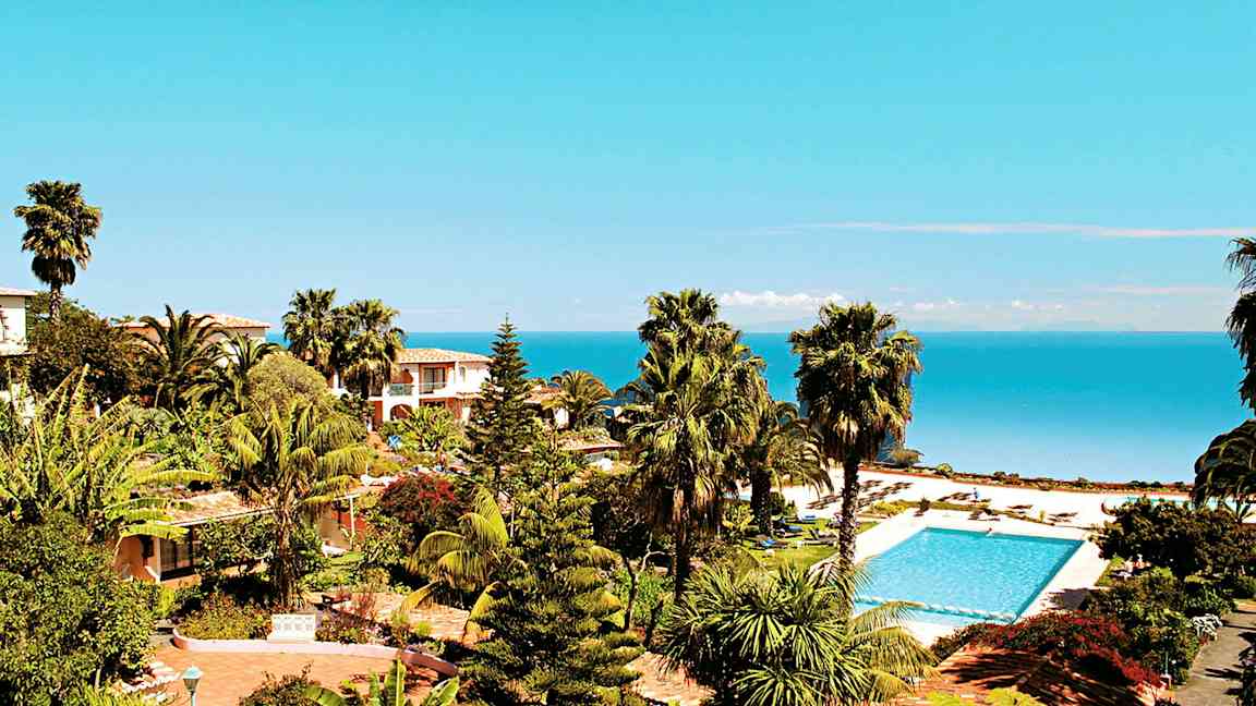 Näkymä trooppiseen puutarhaan, jossa palmuja, uima-allas ja meri taustalla. (Tekoälyn luoma teksti)