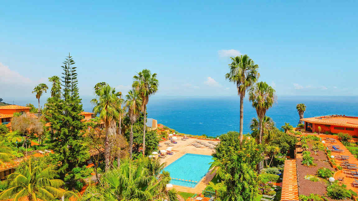 Näkymä uima-allasalueelle, jota ympäröi vehreys ja palmut, taustalla meri. (Tekoälyn luoma teksti)