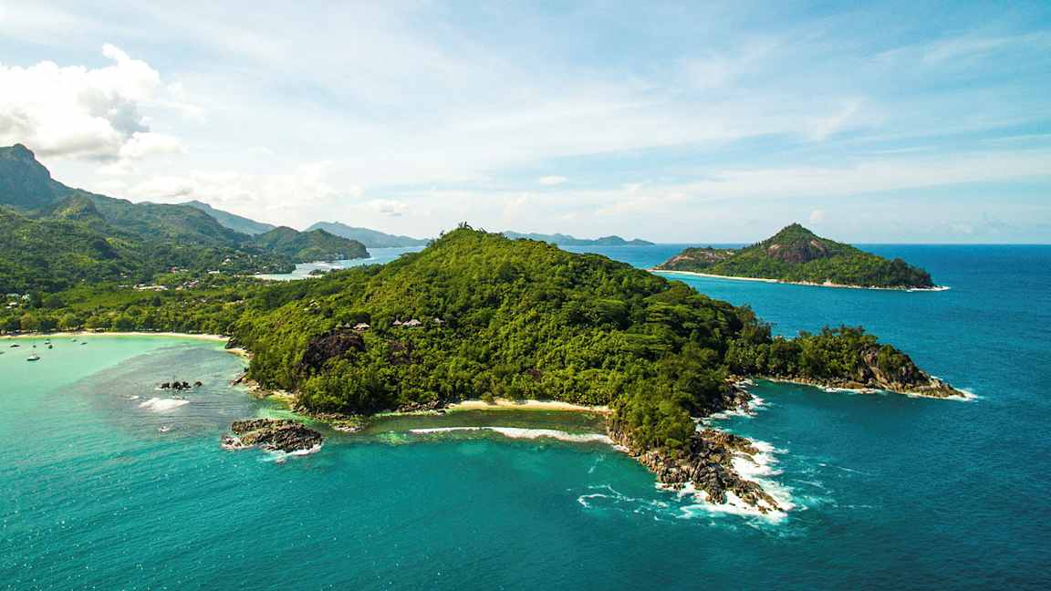 Ilmakuva vehreästä saaresta, jota ympäröi kirkkaan sininen meri ja taustalla näkyy pienempiä saaria. (Tekoälyn luoma teksti)