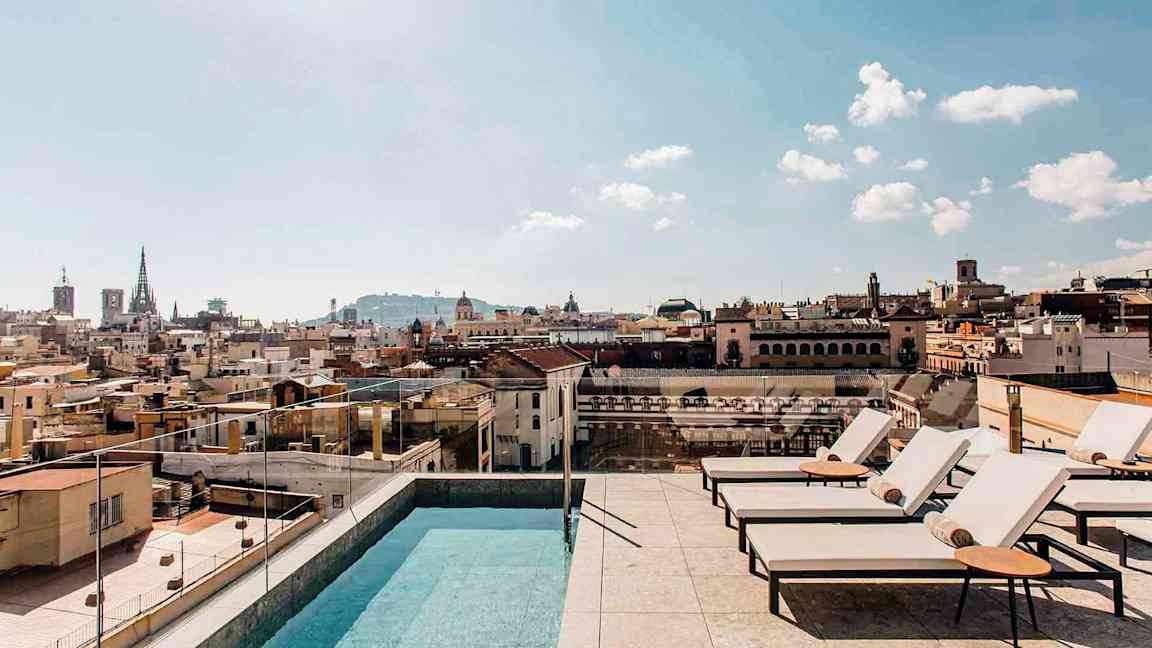 Ulkoallas ja aurinkotuoleja kattoterassilla, josta on näkymä yli kaupungin rakennusten, kirkkaalla taivaalla. (Tekoälyn luoma teksti)