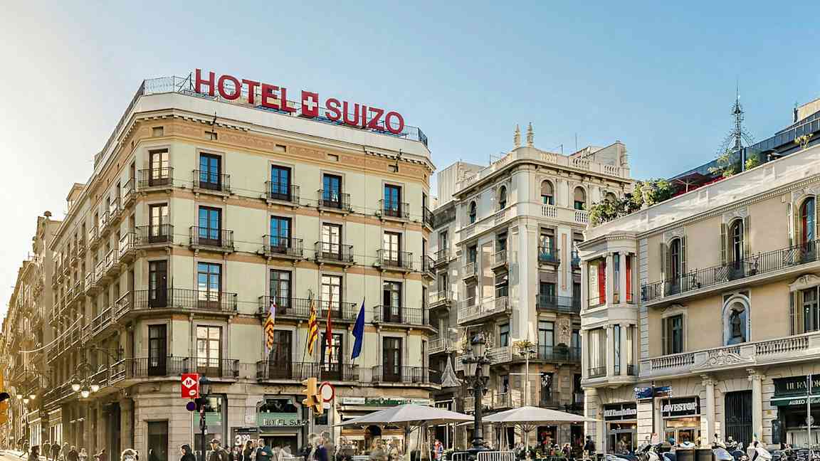 Hotelli nimeltä "Hotel Suizo", joka sijaitsee kulmassa ja jossa on useita kerroksia ja parvekkeita, ympärillä muita rakennuksia ja kadulla kulkevia ihmisiä. (Tekoälyn luoma teksti)