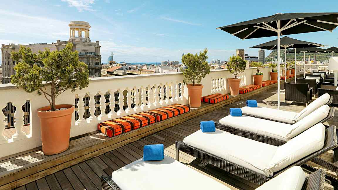 Näköala terassille, jossa aurinkotuoleja ja aurinkovarjoja, taustalla kaupunki ja meri. (Tekoälyn luoma teksti)