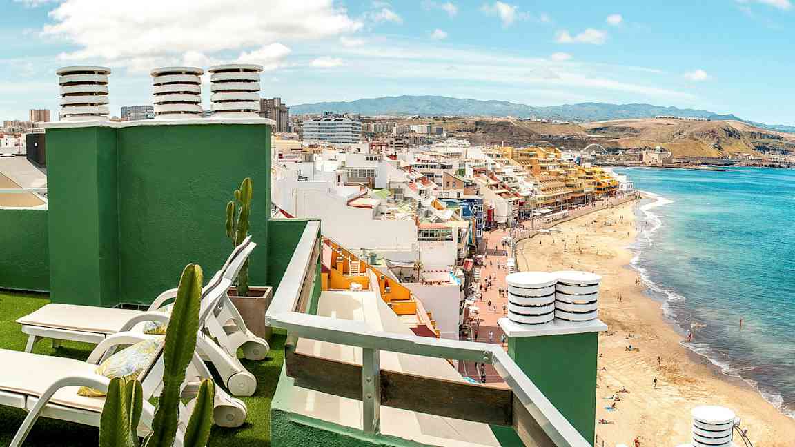 Näkymä kattoterassilta, jossa aurinkotuoleja, kaktuksia ja ranta sekä kaupunki taustalla. (Tekoälyn luoma teksti)