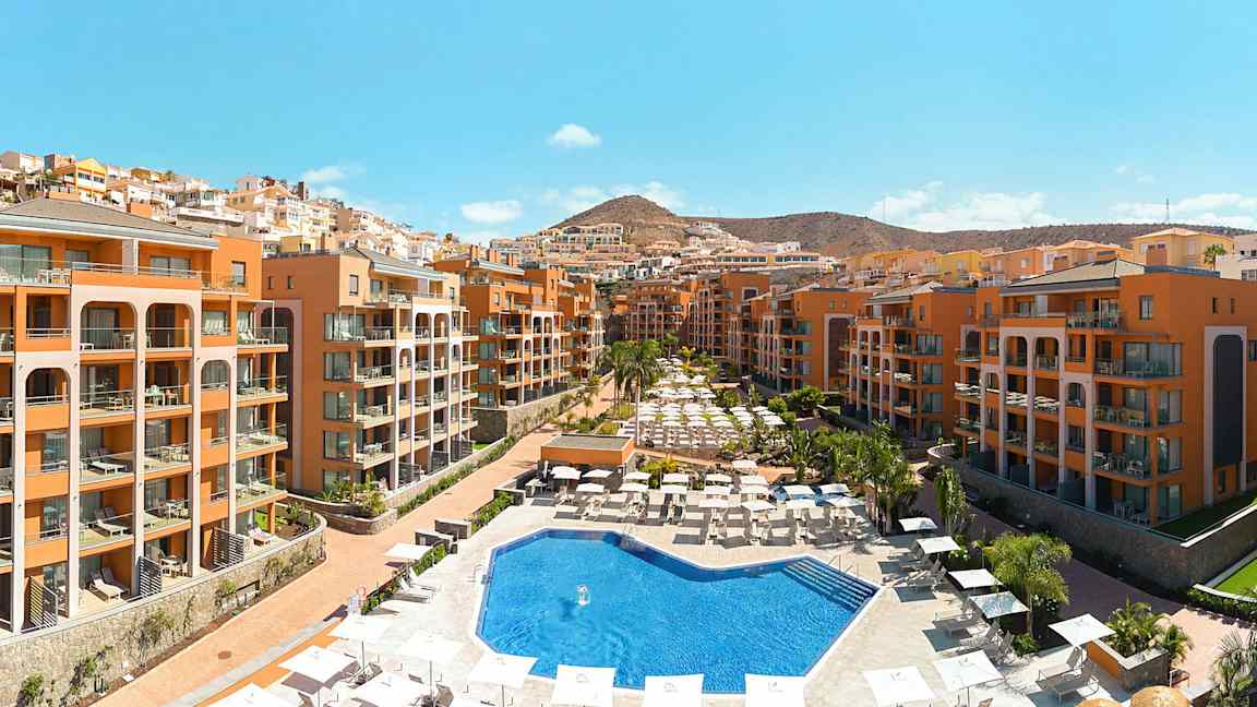 Hotellialue, jossa on ulkouima-allas, ympärillä aurinkotuoleja ja -varjoja, sekä rakennuksia terrakotan värissä. (Tekoälyn luoma teksti)