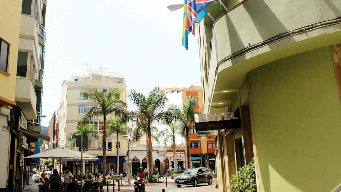 Katu, jolla on rakennuksia, palmuja ja hotellikyltti. Ihmiset istuvat kahvilassa ja moottoriajoneuvo ajaa ohi. (Tekoälyn luoma teksti)