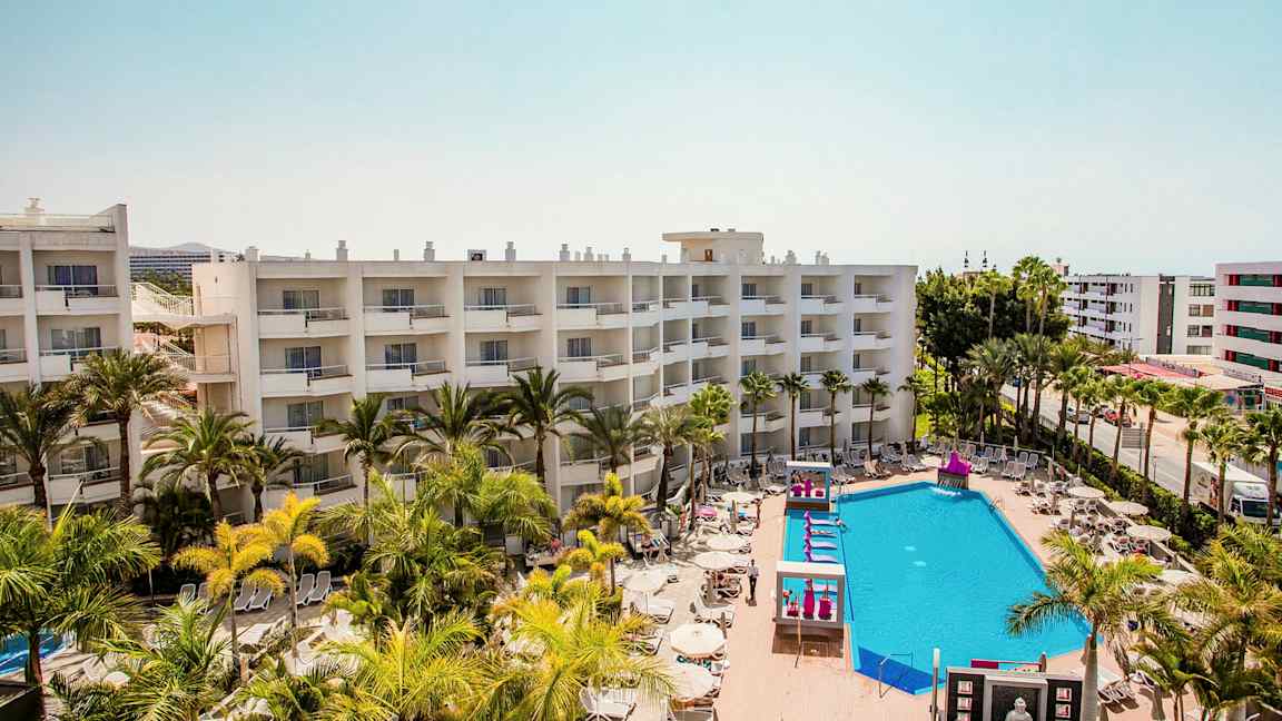 Hotellirakennukset parvekkeineen, uima-allasalue aurinkotuoleineen ja varjon alle palmuineen. (Tekoälyn luoma teksti)