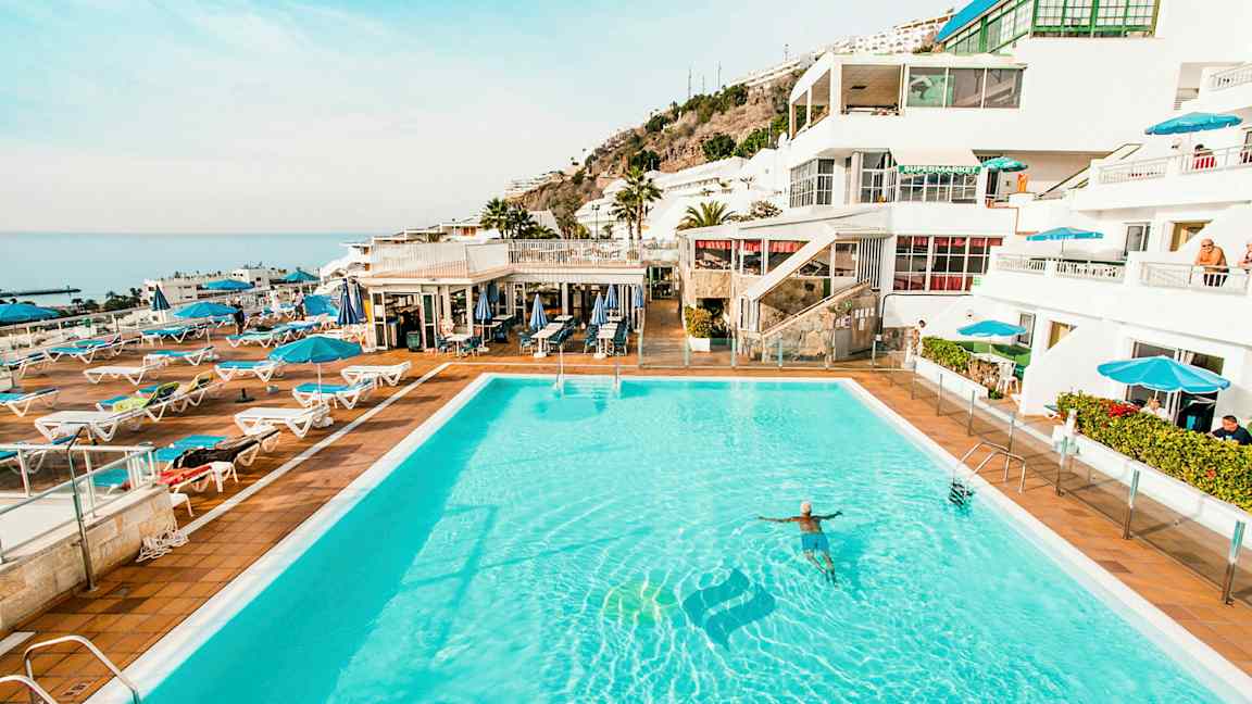 Kuva suuresta ulkouima-altaasta, jota ympäröivät aurinkotuolit ja aurinkovarjot, taustalla valkoisia rakennuksia ja meri. (Tekoälyn luoma teksti)