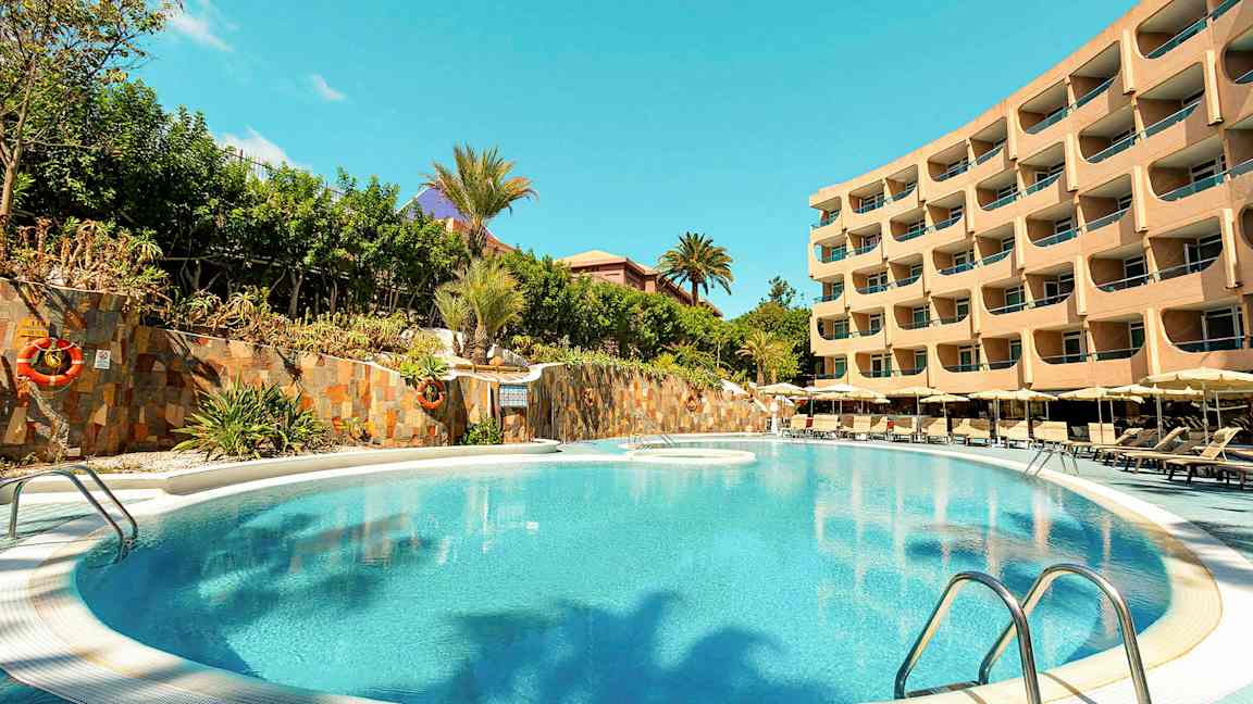 Kuva uima-altaasta, jonka ympärillä on aurinkotuoleja ja taustalla monikerroksinen hotellirakennus. (Tekoälyn luoma teksti)