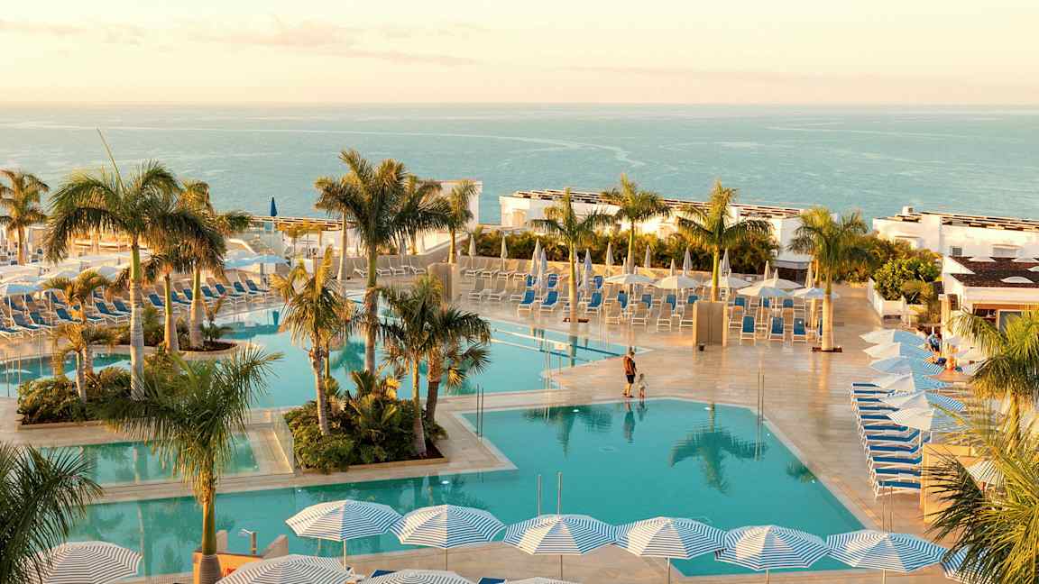 Uima-allas aurinkotuoleilla ja aurinkovarjoilla, ympärillä palmuja ja taustalla meri. (Tekoälyn luoma teksti)