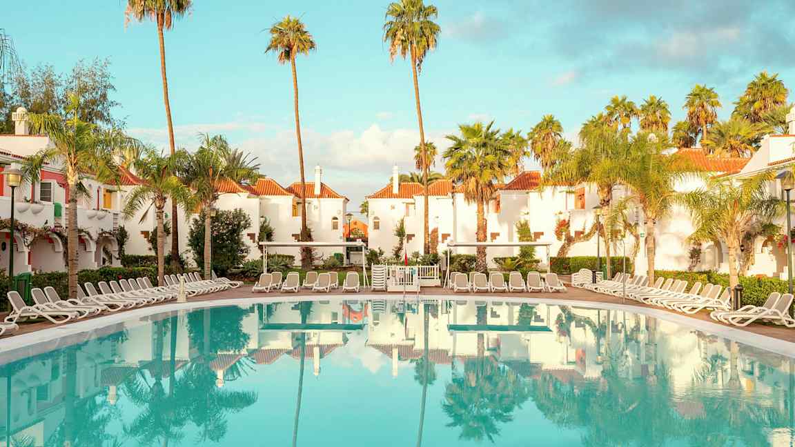 Hotellialue, jossa on valkoiseksi rapattuja rakennuksia punaisilla katoilla, ympäröity palmuilla, ja suuri uima-allas aurinkotuoleilla. (Tekoälyn luoma teksti)