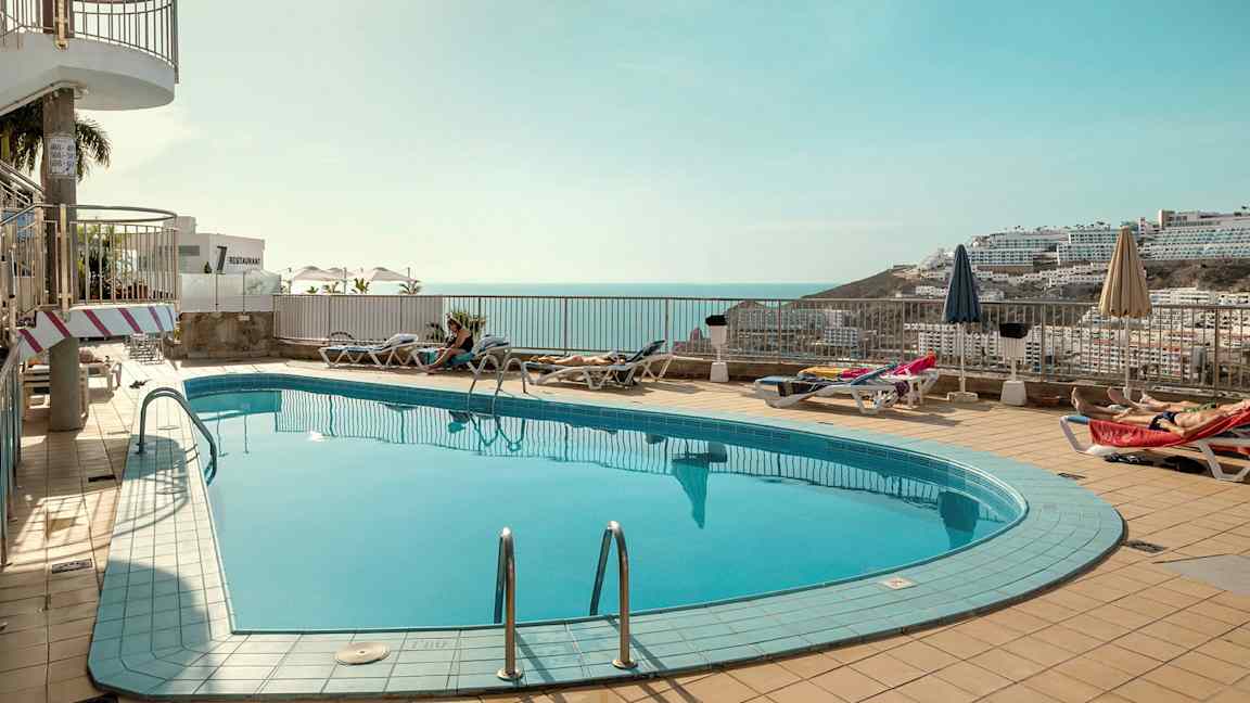 Ulkouima-allas, jota ympäröivät aurinkotuolit ja aurinkovarjot, taustalla merinäköala ja ympäröivät rakennukset. (Tekoälyn luoma teksti)