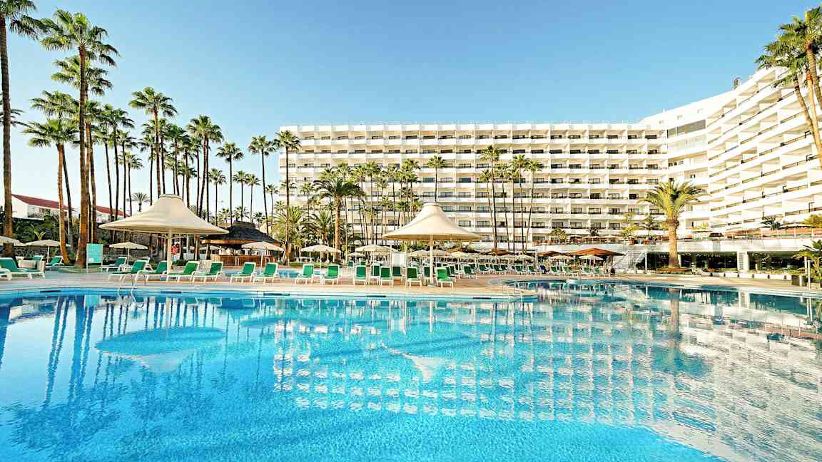 Hotellirakennus, jossa suuri uima-allas ja aurinkotuoleja etualalla, ympäröitynä palmuilla. (Tekoälyn luoma teksti)