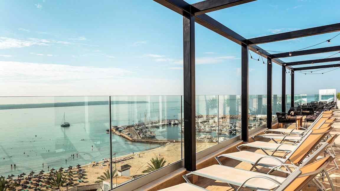 Aurinkotuoleja asetettu lasikaiteen eteen, näkymä rannalle ja satamaan. (Tekoälyn luoma teksti)