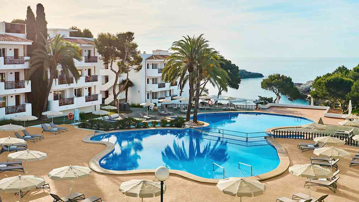 Hotellialueen kuva, jossa on suuri uima-allas, jota ympäröivät aurinkotuolit ja -varjot. Taustalla näkyy meri ja osia rakennuksesta. (Tekoälyn luoma teksti)