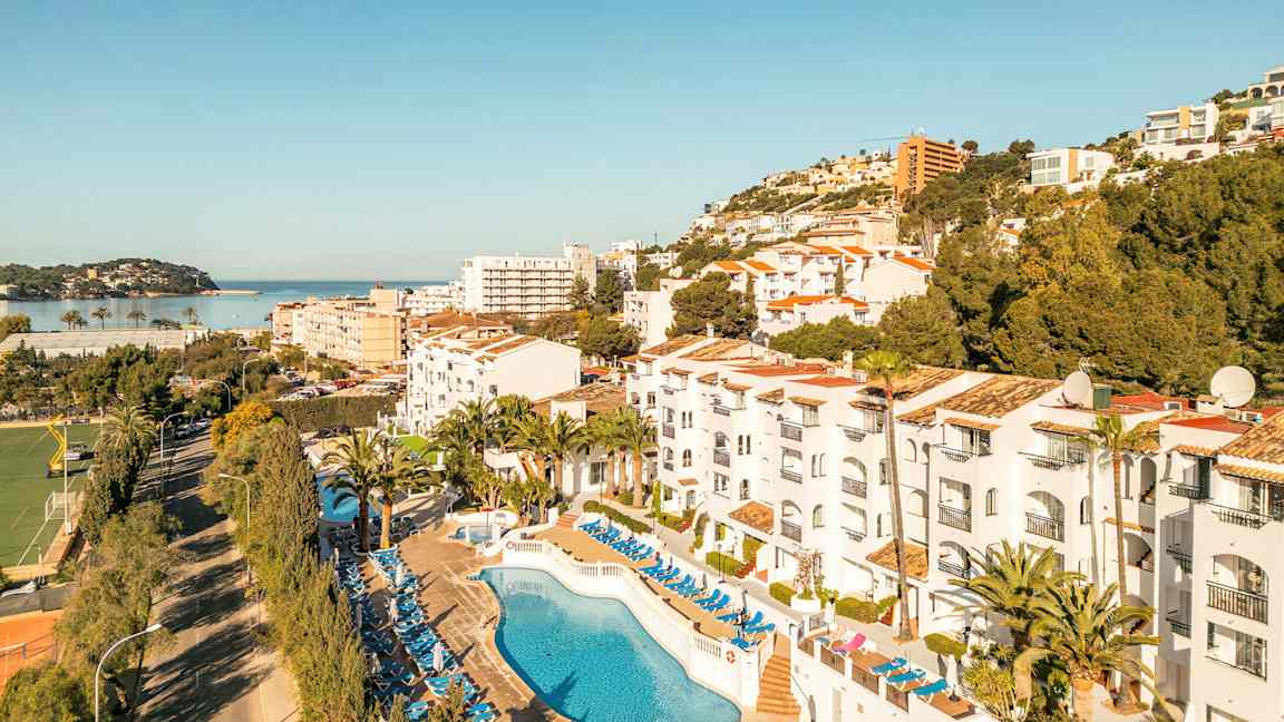 Kuva hotellialueesta, jossa on uima-allas ja aurinkotuoleja, ympäröivänä puita ja rakennuksia sekä näkymä merelle. (Tekoälyn luoma teksti)