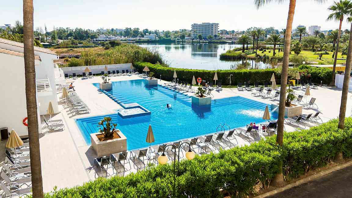 Ulkoallas aurinkotuoleineen ja aurinkovarjoineen, ympärillä vihreitä kasveja ja näkymä järvelle ja avoimeen maisemaan. (Tekoälyn luoma teksti)