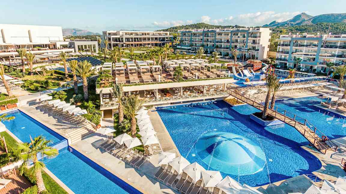 Näkymä hotellialueelle, jossa on uima-altaita, aurinkotuoleja ja rakennuksia. (Tekoälyn luoma teksti)