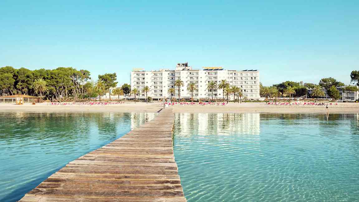 Puusilta johtaa hiekkarannalle, taustalla hotellirakennuksia ja palmuja. (Tekoälyn luoma teksti)