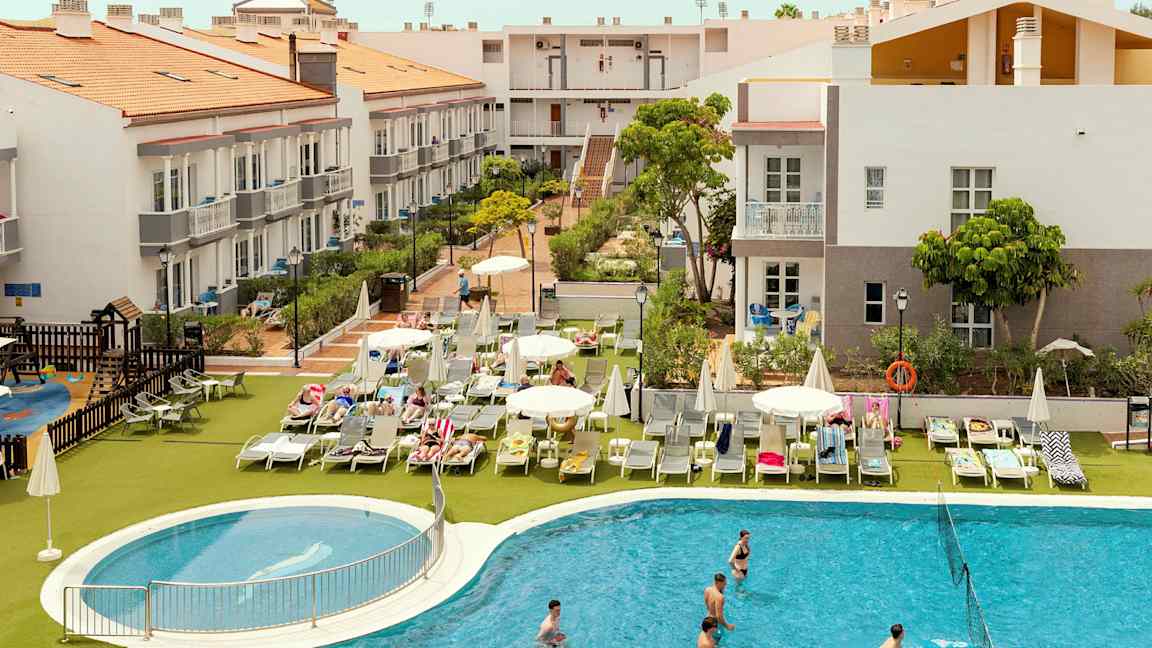 Hotellialue, jossa rakennuksia, aurinkotuoleja ja uima-allas. (Tekoälyn luoma teksti)