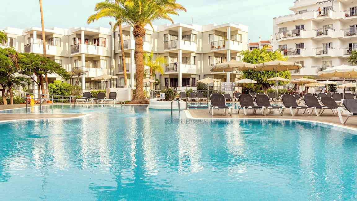 Ulkoallas, jonka ympärillä on aurinkotuoleja ja aurinkovarjoja, taustalla nykyaikainen rakennus. (Tekoälyn luoma teksti)