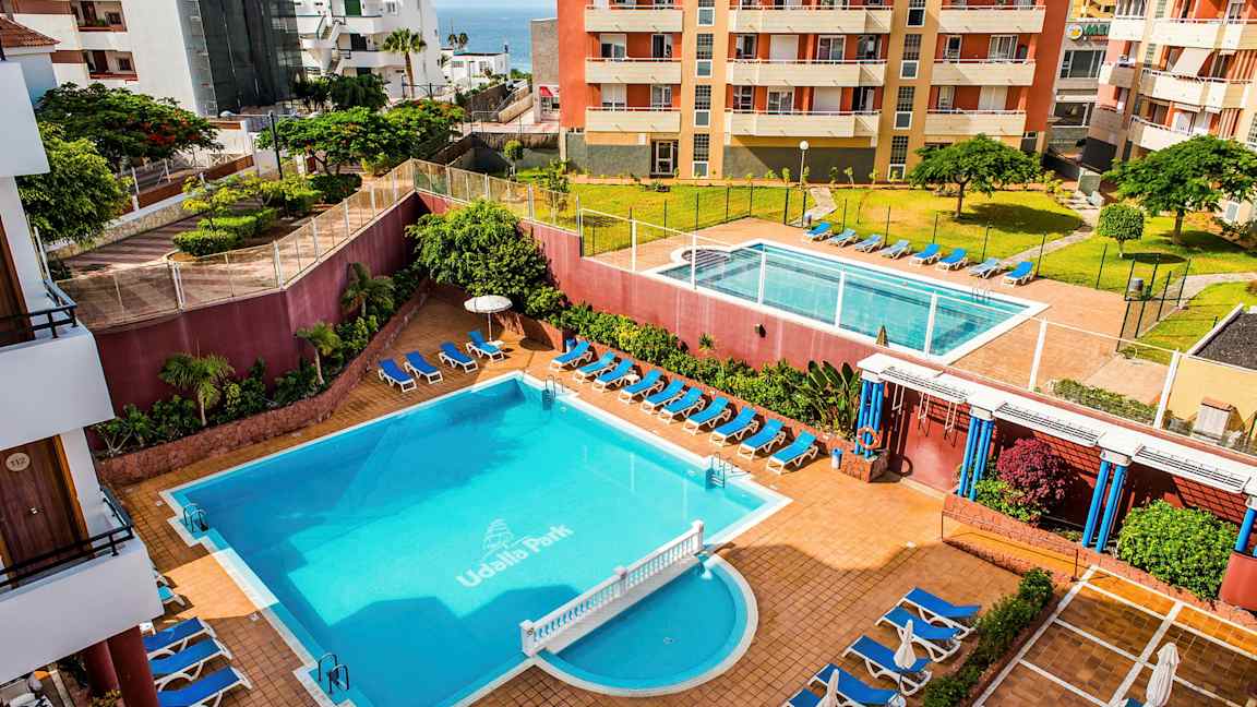 Hotellialue, jossa on kaksi uima-allasta, aurinkotuoleja ja ympäröiviä rakennuksia. (Tekoälyn luoma teksti)