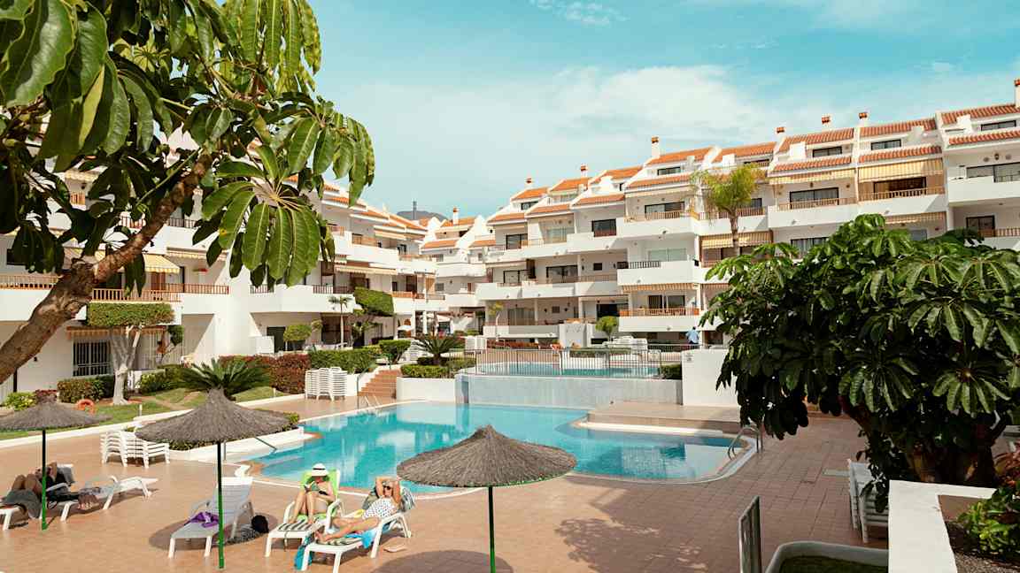 Ulkotila, jossa on uima-allas, aurinkotuoleja ja monikerroksinen hotelli. Useita aurinkovarjoja ja vihreitä kasveja ympäröivät aluetta. (Tekoälyn luoma teksti)