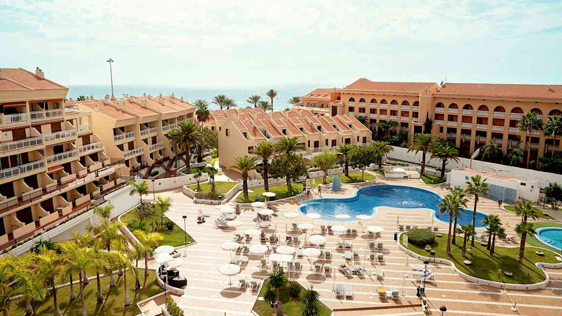 Useat terrakottakattoiset rakennukset ympäröivät uima-allasta ja aurinkotuoleja, ympärillä palmuja ja meri taustalla. (Tekoälyn luoma teksti)