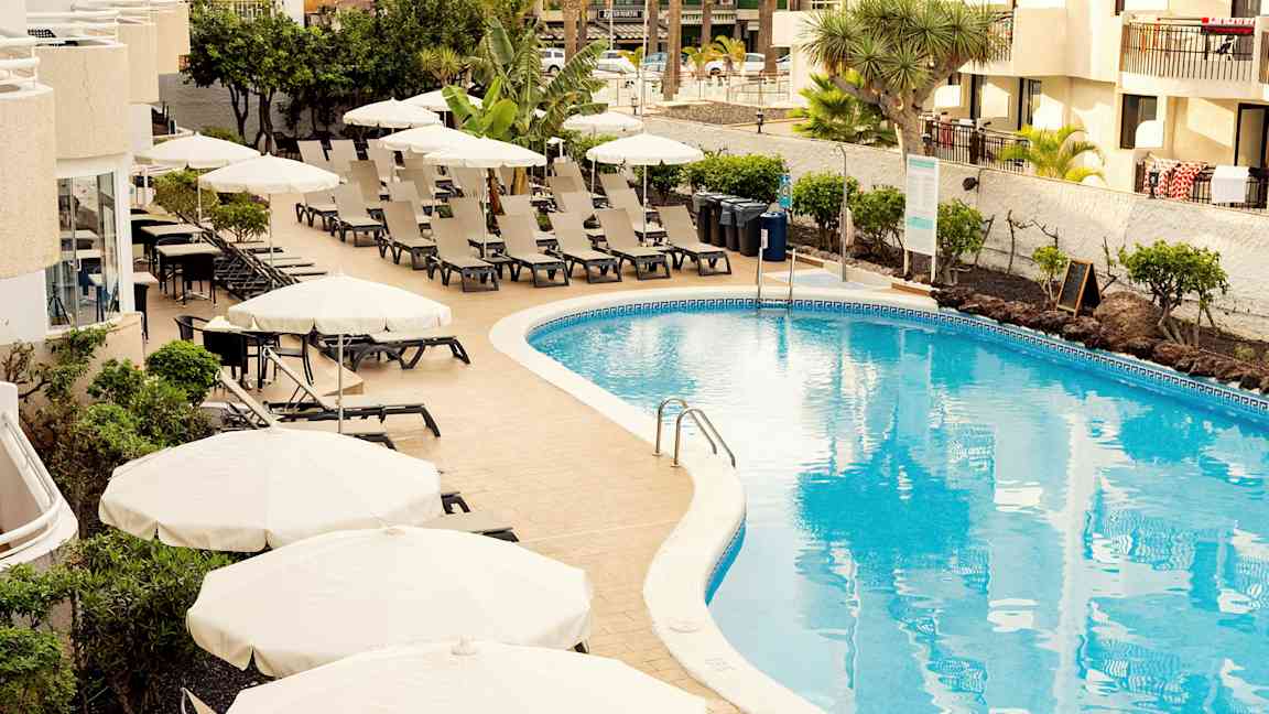 Uima-allasalue aurinkotuoleilla ja varjoilla hotellin vieressä. (Tekoälyn luoma teksti)