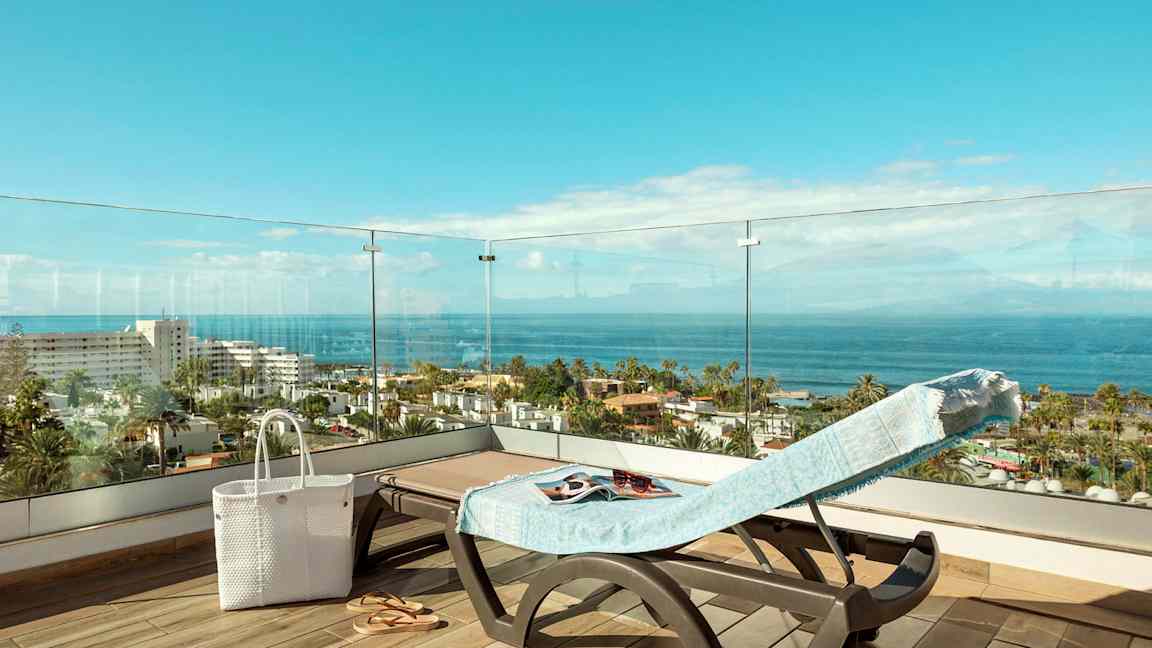 Näkymä terassilta aurinkotuolille, jossa on pyyhe, valkoinen laukku ja sandaalit, näkyy meri ja rakennuksia. (Tekoälyn luoma teksti)