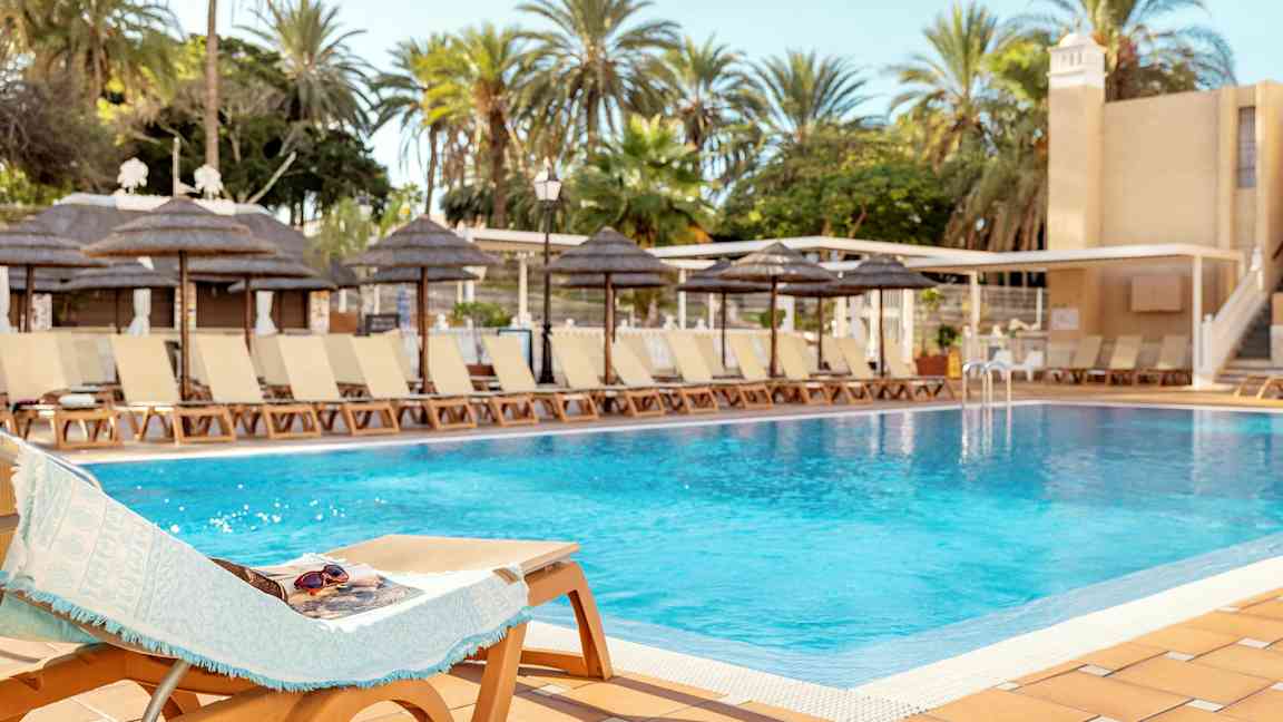 Uima-allas aurinkotuolien ja palmujen ympäröimänä kirkkaan taivaan alla. (Tekoälyn luoma teksti)