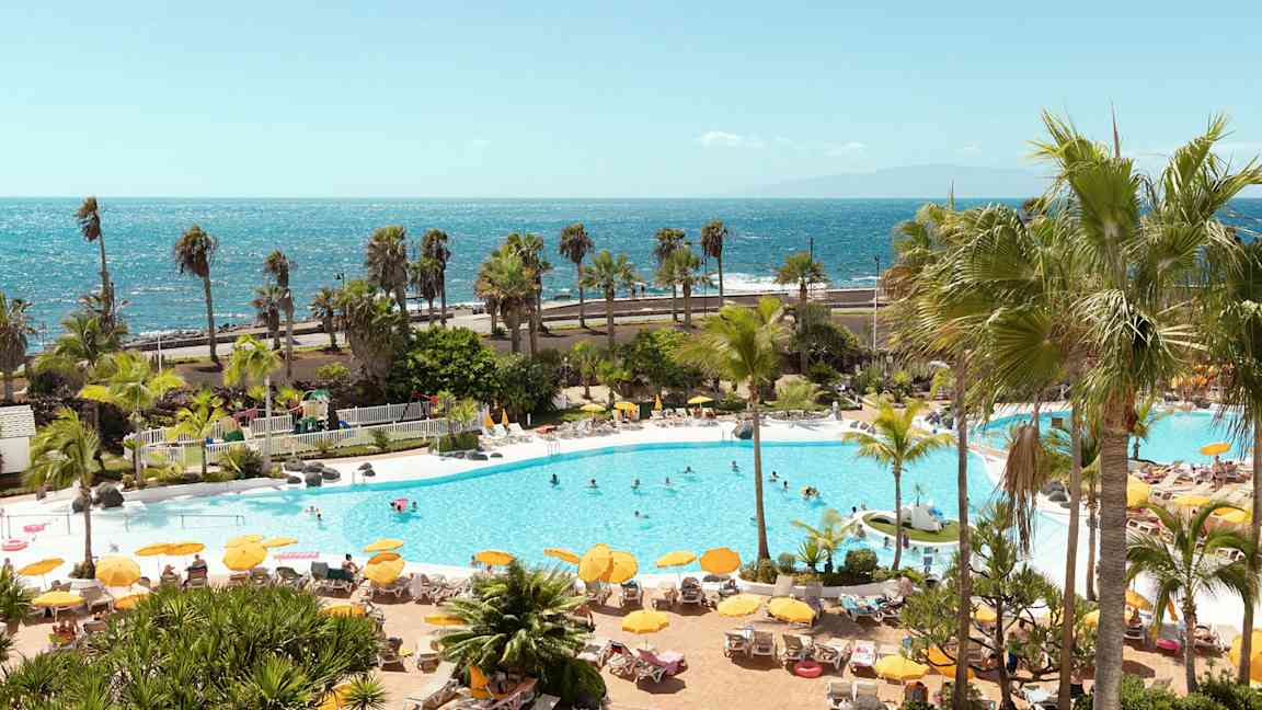 Uima-allasalue, jossa aurinkotuoleja, aurinkovarjoja ja ihmisiä, taustalla meri. (Tekoälyn luoma teksti)